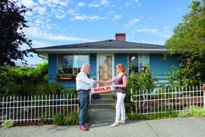 An older man and a younger woman shake hands over a "Sold" sign to celebrate a successful home sale.