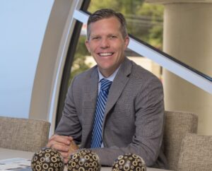 A photo of Adam Kenney, where he poses seated at a desk in front of a window.